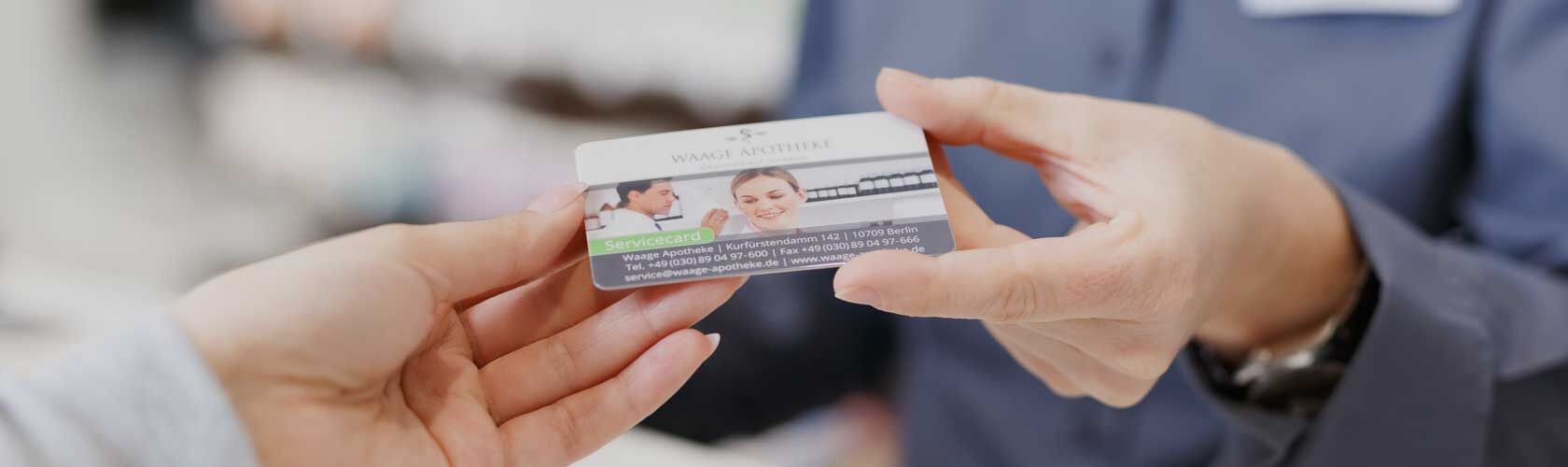 Die Kundekarte der Waage Apotheke Eine Person überreicht einer anderen eine Servicekarte, die Informationen und ein Bild von einem lächelnden Paar zeigt.